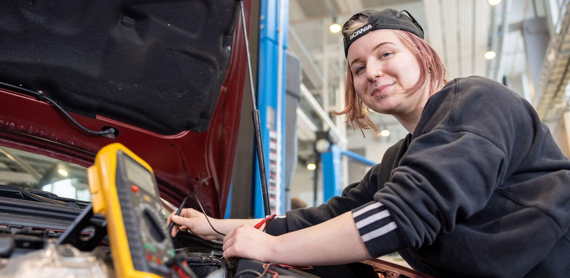 Henkilö mittaa jotakin autosta. Auton konepelti on auki.