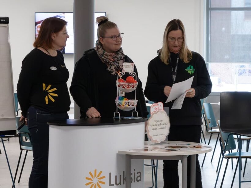 Kolme henkilöä seisoo aulassa ja ottaa vastaan vieraita.