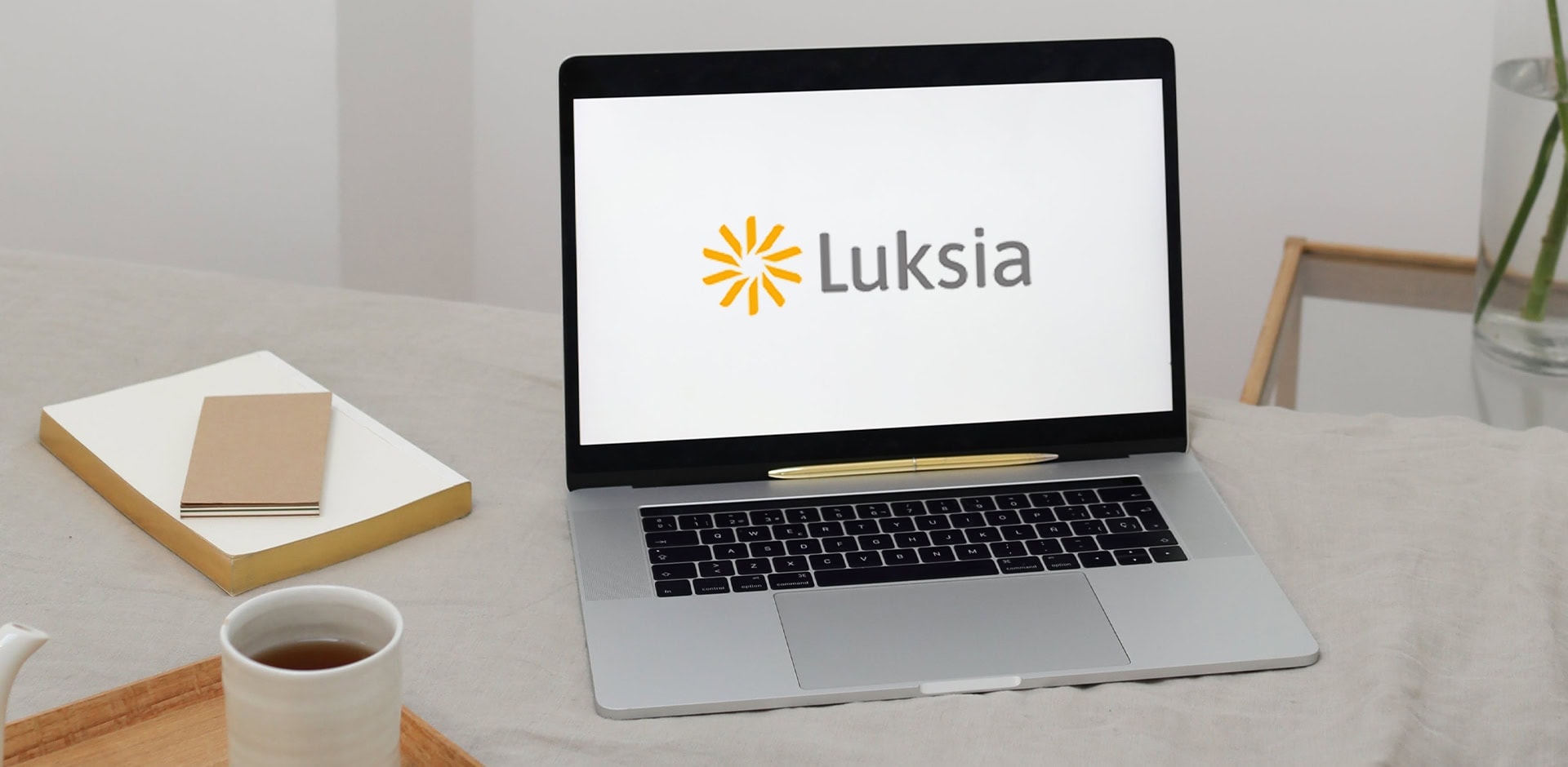 Pöydällä on kannettava, jossa on näytöllä Luksian logo, kirjoja sekä teekuppi.