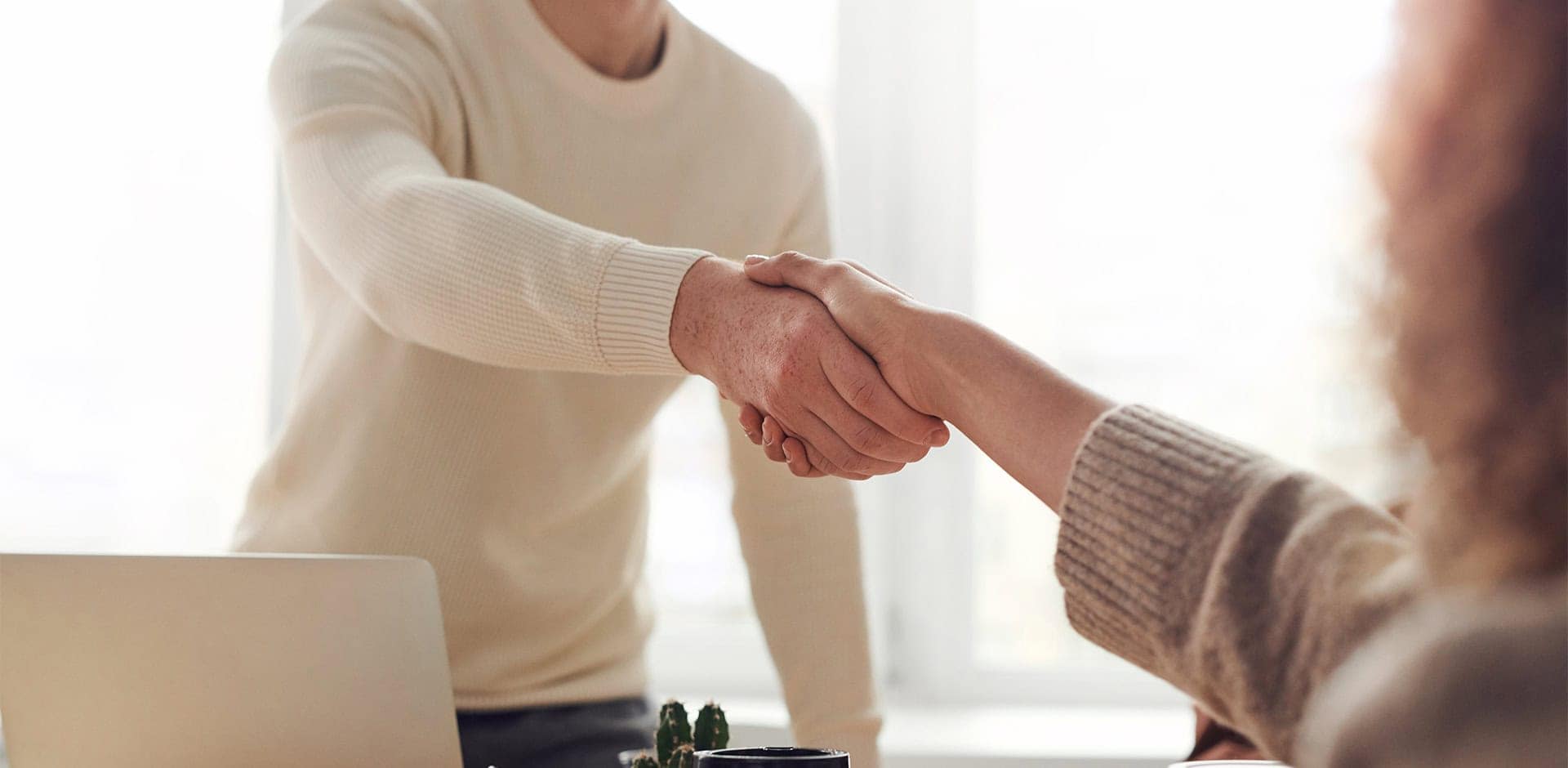 Kaksi ihmistä kättelee pöydän yli. Toisella on valkoinen paita ja toisen henkilön paita on beige.
