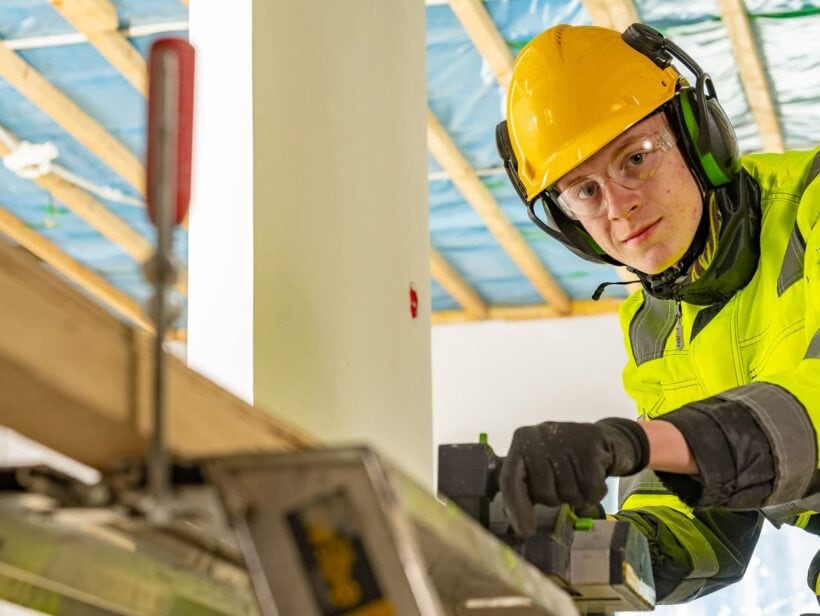 Henkilö työvaatteissa käsittelee puuta rakennustyömaalla.