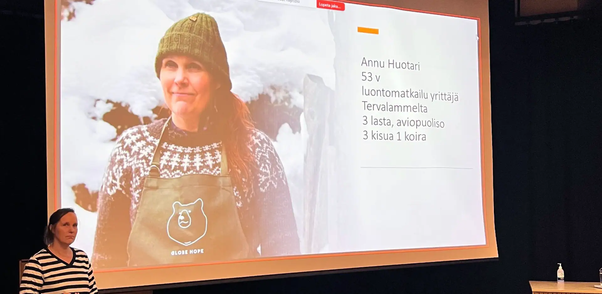 Lavalla henkilö pitää puhetta. Taustalla kuva henkilöstä, joka on lumisessa maisemassa villapaita ja pipo päässä.