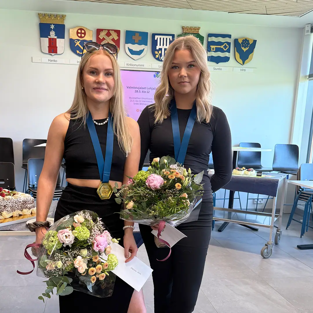 Kaksi vaaleahiuksista henkilöä mustissa vaatteissa, mitalit kaulassa ja värikäs kukkakimppu kädessä.