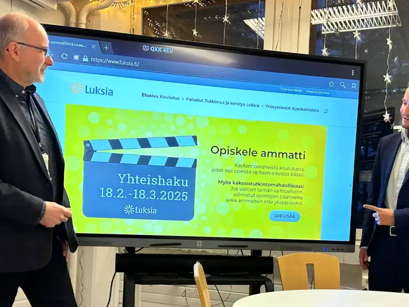 Kaksi miestä seisoo ison TV näytön vieressä ja tutkivat kuvassa olevaa luksia.fi etusivua. Sivulla on iso keltainen banneri.