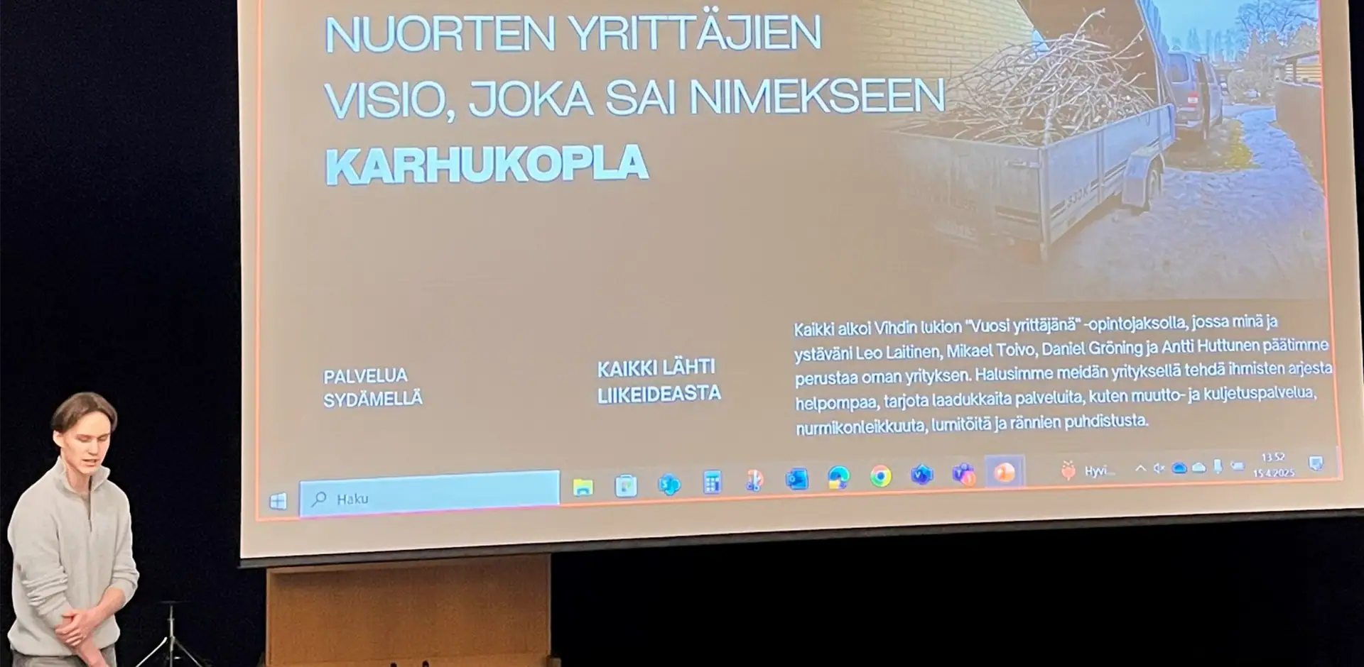 Henkilö pitää esitystä lavalla, jonne on heijastettu tekstiä, jossa lukee Karhukopla.