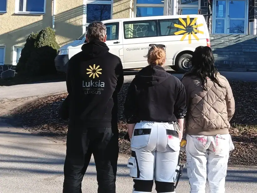 Kolme henkilöä selin kameraan kampuksen pihalla, jossa valkoinen pakettiauto ja rakennuksen julkisivu.