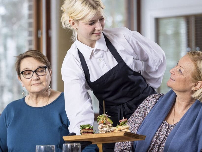 Kolme henkilöä ravintolassa. Kaksi asiakasta ja tarjoilija.