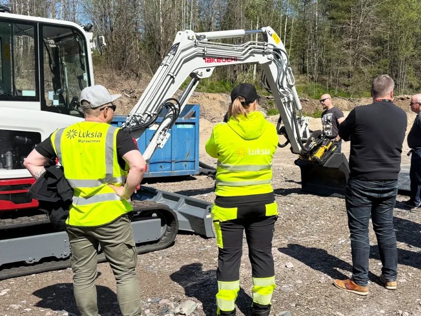 Valkoinen kaivinkone, jossa punaista. Ympärillä ihmisiä.