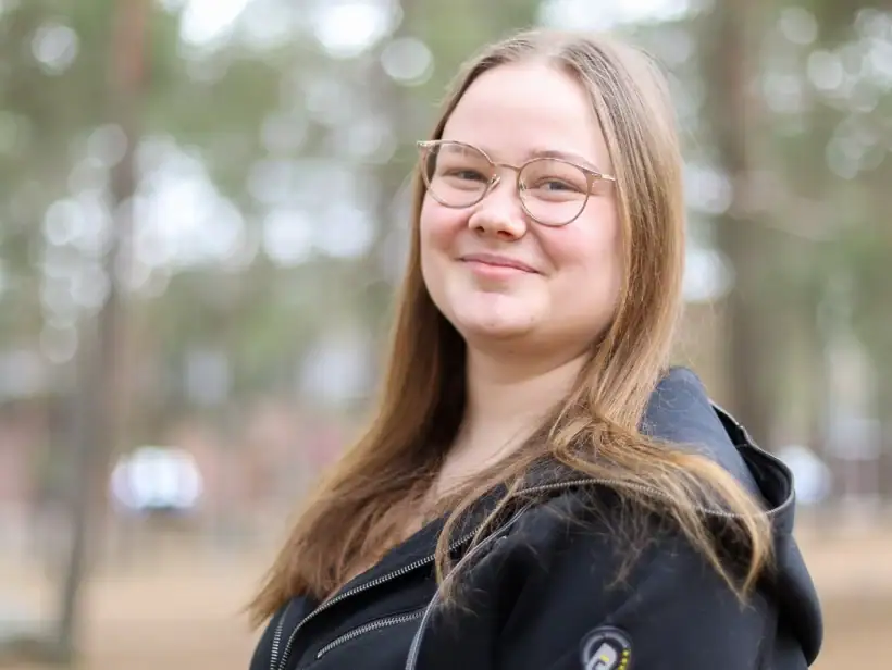 Henkilö mustassa hupparissa seisoo puiston laidalla ja hymyilee kameraan.