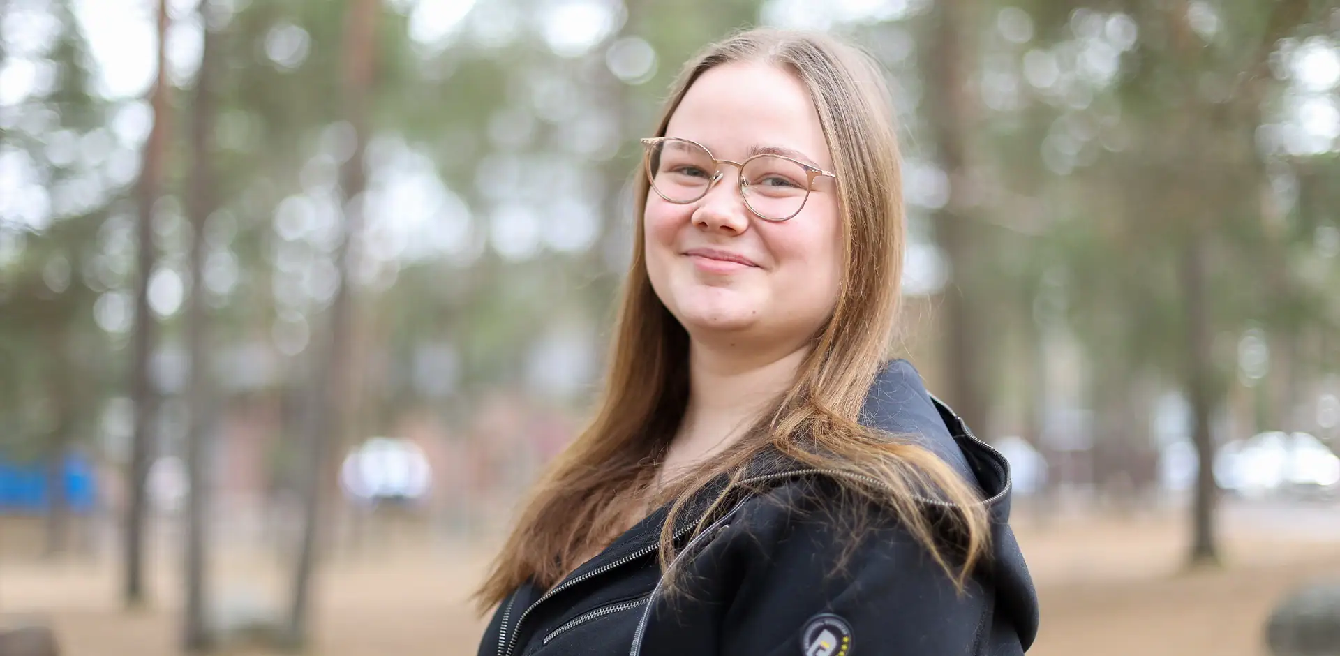 Henkilö mustassa hupparissa seisoo puiston laidalla ja hymyilee kameraan.