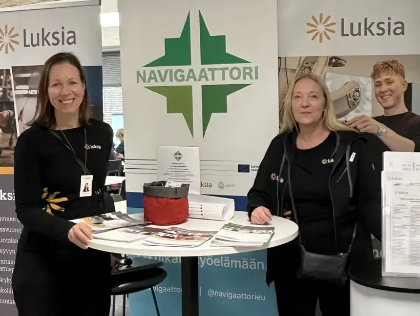 Kaksi henkilöä mustissa vaatteissa seisovat Navigaattorilogollisen taustan edessä ja hymyilevät kameralle.