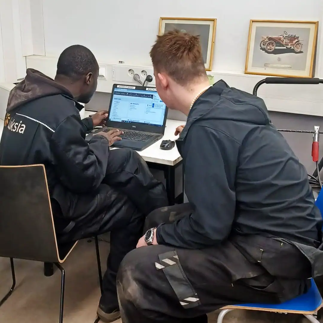 Kaksi henkilöä tummissa vaatteissa istuu ja katsoo työpöydällä olevaa päätettä.