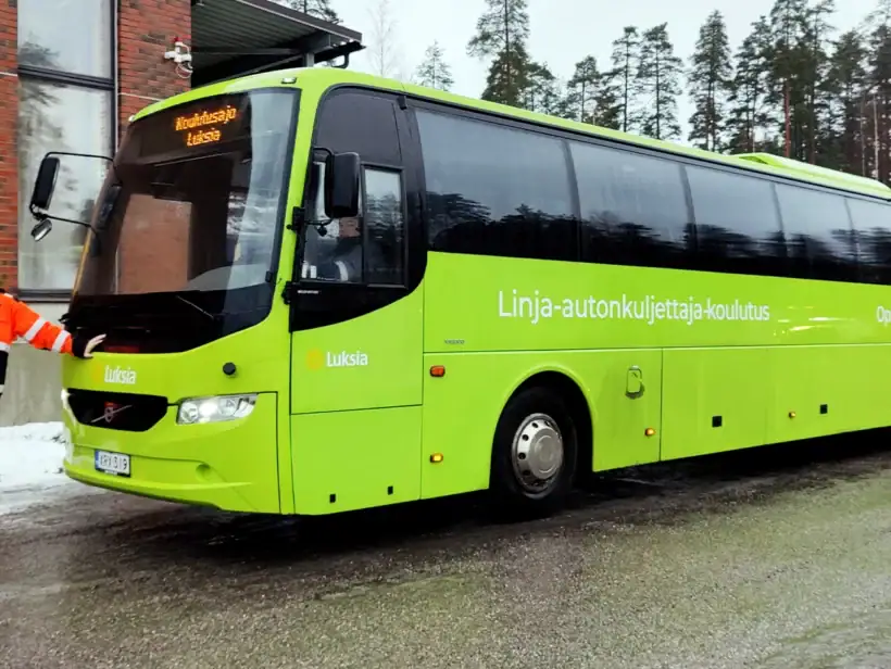 Vihreä linja-auto talvisessa maisemassa. Auton keulaan nojaa henkilö oranssimustissa huomiovaatteissa.