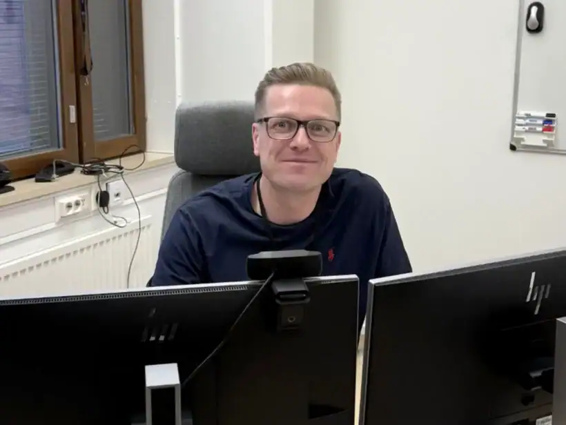 Henkilö mustassa paidassa istuu työpöydän takana. Pöydällä kaksi mustaa näyttöä.