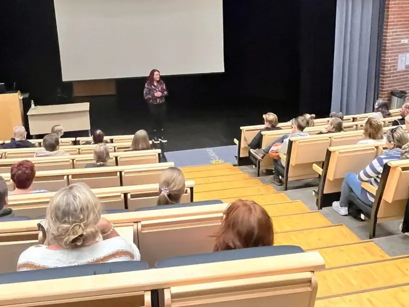 Kampuksen auditoriossa on yleisöä sekä lavalla on puhuja, jolla on mustat housut ja värikäs paita.