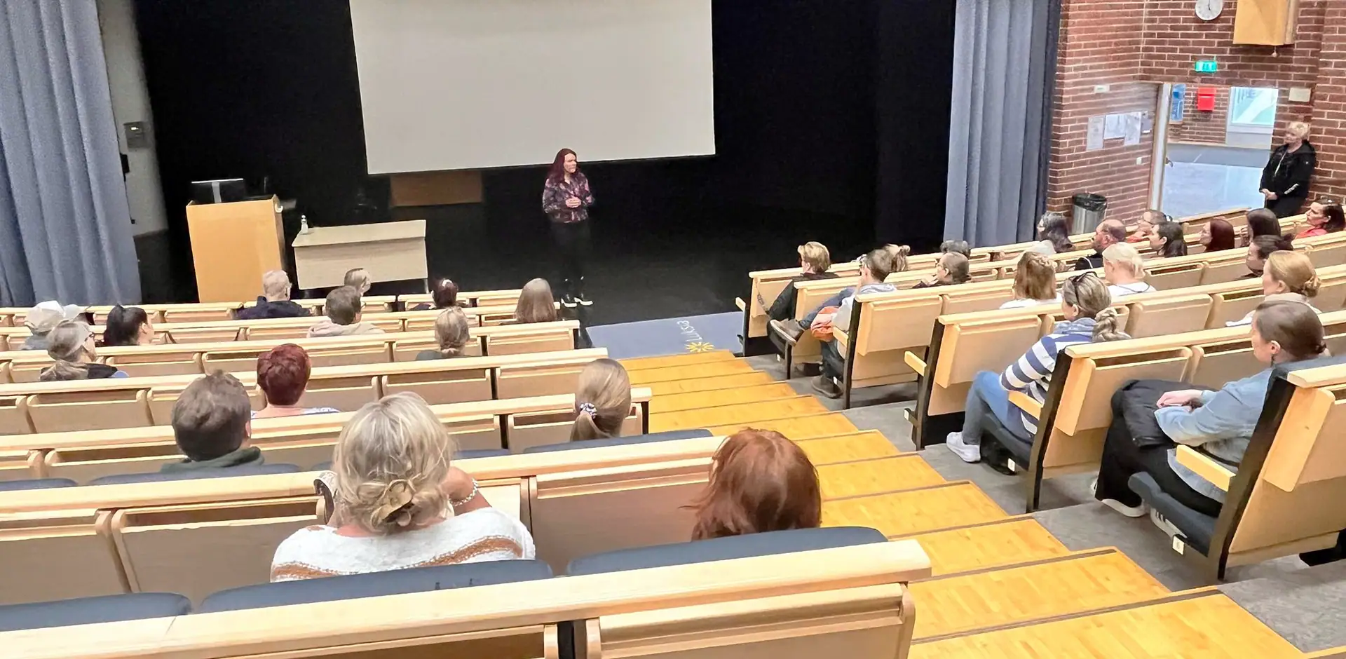 Kampuksen auditoriossa on yleisöä sekä lavalla on puhuja, jolla on mustat housut ja värikäs paita.