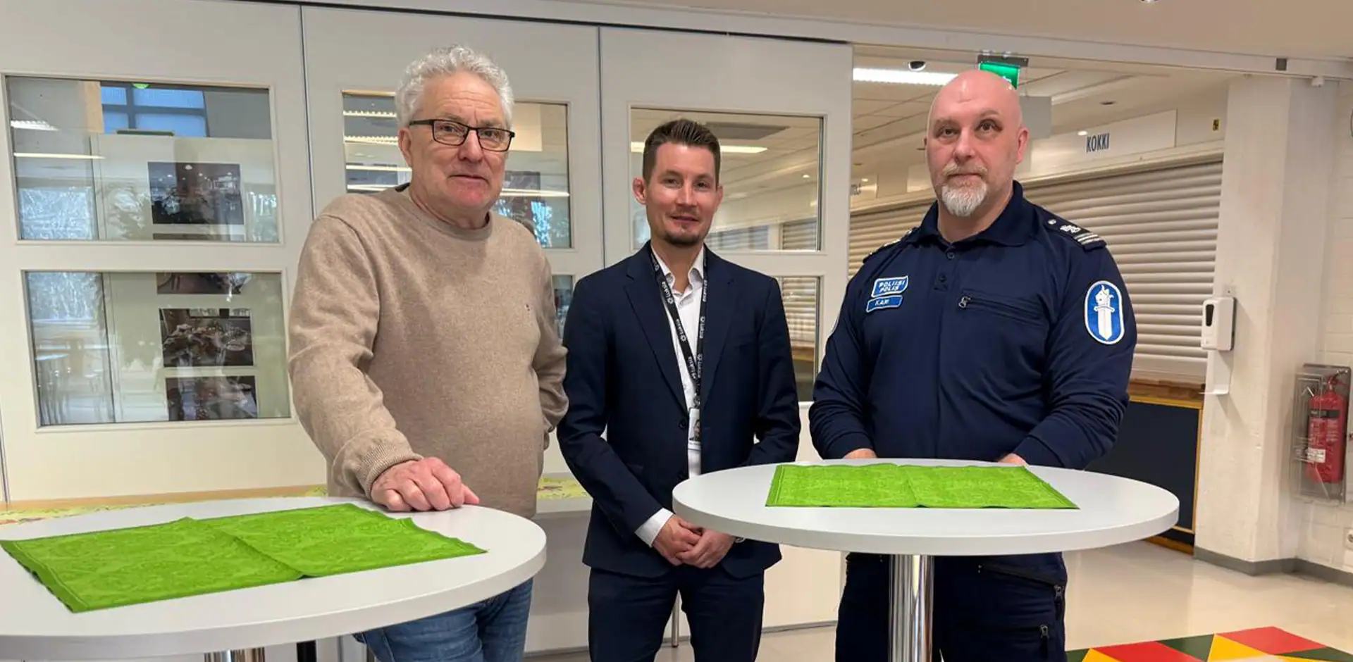 Kolme henkilöä ruokalamiljöössä. Yksi henkilöistä on poliisin virka-asussa.