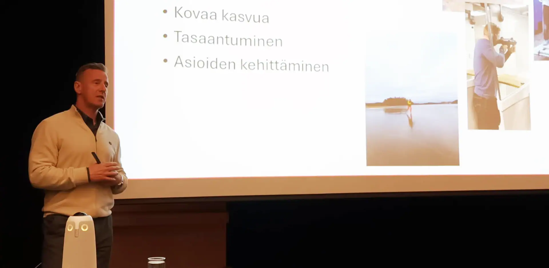 Henkilö esiintyy lavalla, jonne on heijastettu kuva, jossa tekstiä ja kuvia.