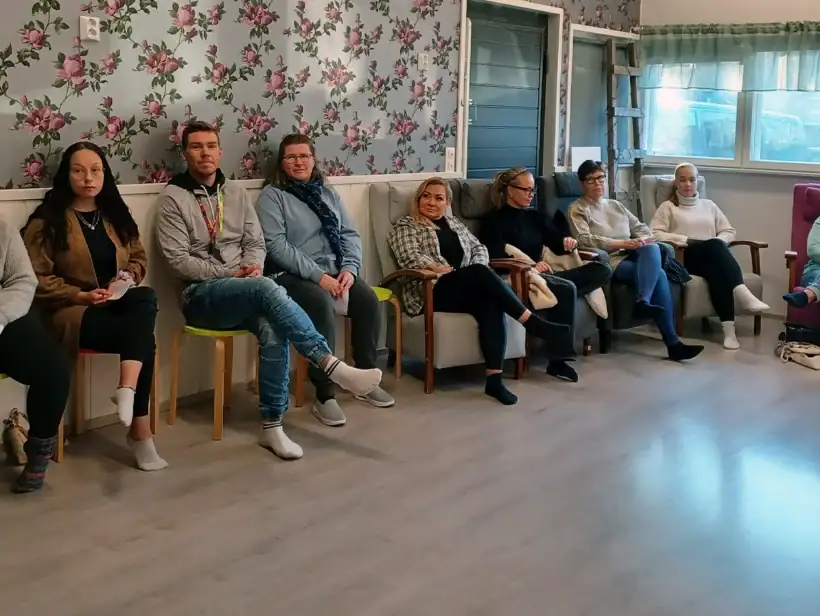 Joukko ihmisiä istuu kukkatapetilla tapetoidussa tilassa. Tuolit ovat aseteltu seinustalle.