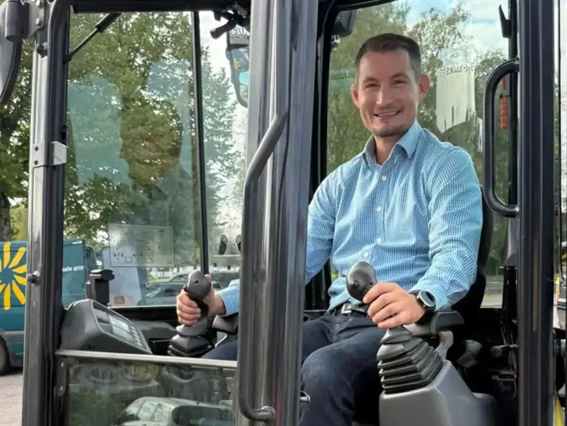 Henkilö sinisessä paidassa istuu keltaisen kaivinkoneen ohjaamossa.