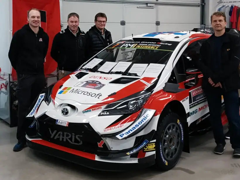 Toyota merkkisen, punavalkoisen ralliauton ympärillä seisoo joukko ihmisiä ja katsovat kohti kameraa.
