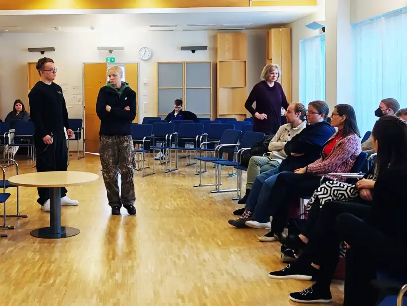 Luksian opiskelijat antavat turvallisuusalan koulutusta yrityksen henkilökunnalle tilassa, jossa on musta piano ja sinisiä tuoleja.