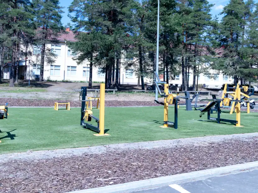 Vihreällä kentällä, koulurakennuksen lähellä, keltaisia ulkokuntosalilaitteita.