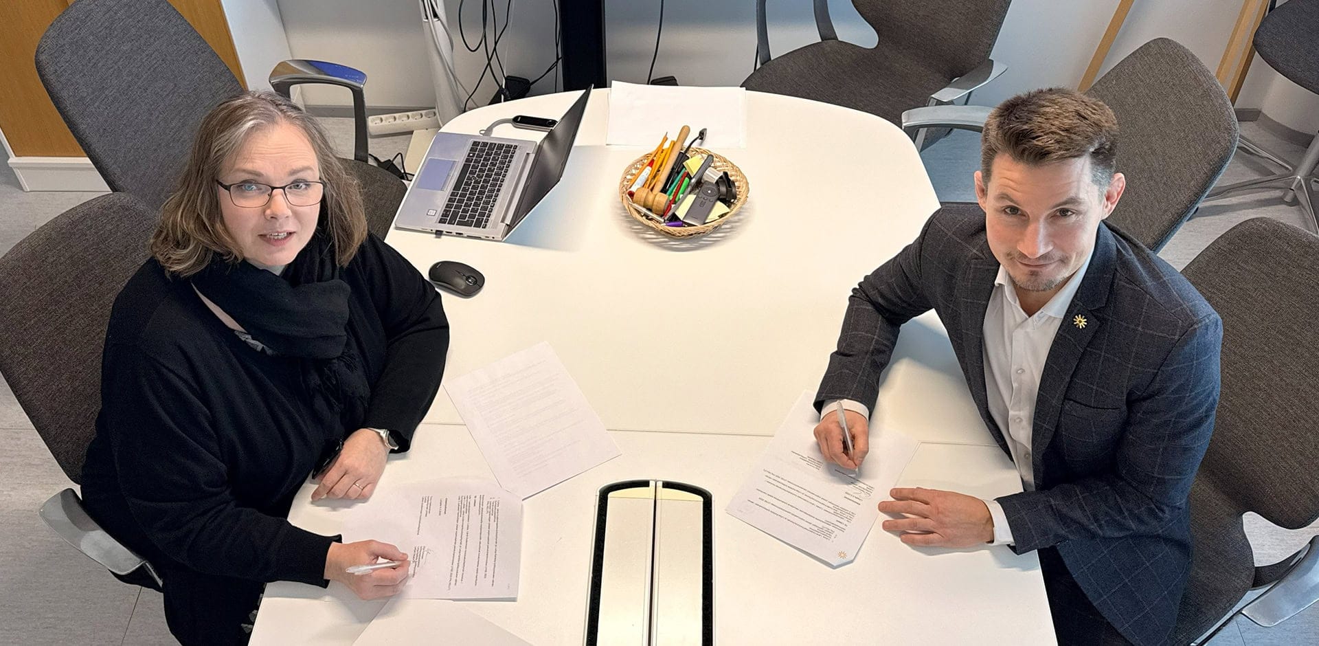 Kaksi henkilöä pöydän ääressä kirjoittamassa asiakirjaan allekirjoituksia.
