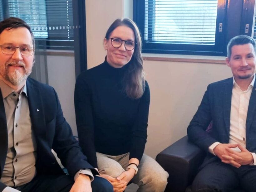 Kolme henkilöä tummissa vaatteissa istuvat ja katsovat kameraan.