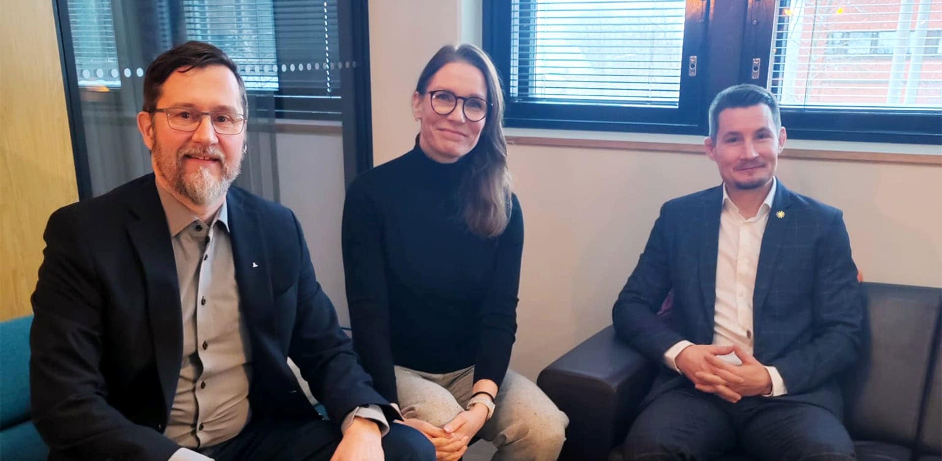 Kolme henkilöä tummissa vaatteissa istuvat ja katsovat kameraan.