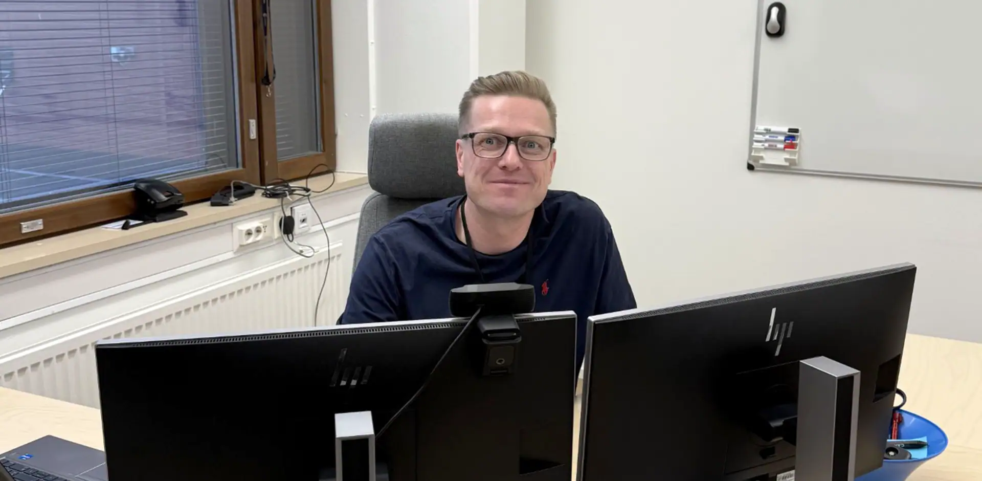 Henkilö mustassa paidassa istuu työpöydän takana. Pöydällä kaksi mustaa näyttöä.