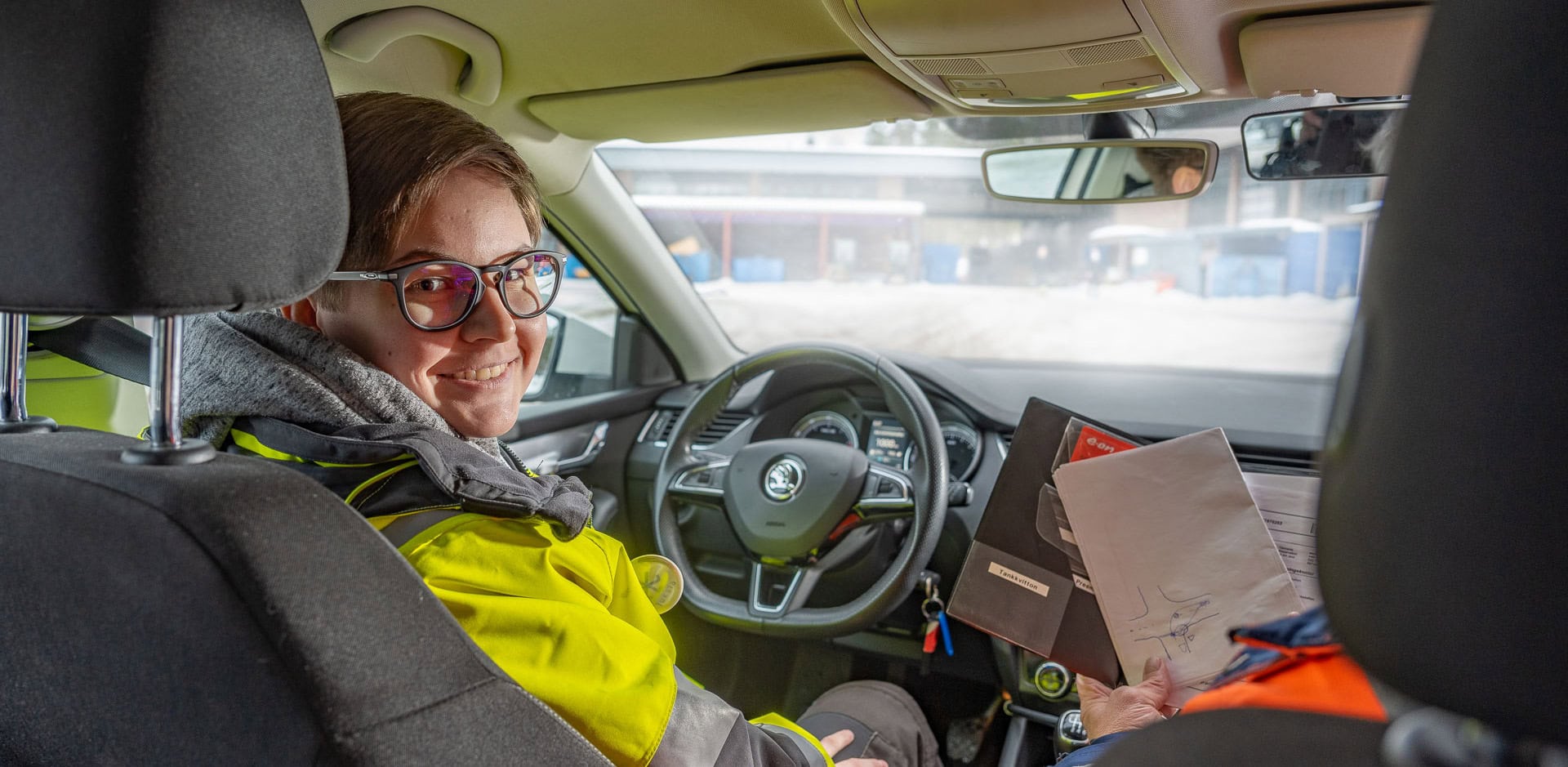 Henkilö istuu autossa kuskin paikalla ja hymyilee kuvaajalle, joka istuu takapenkillä.