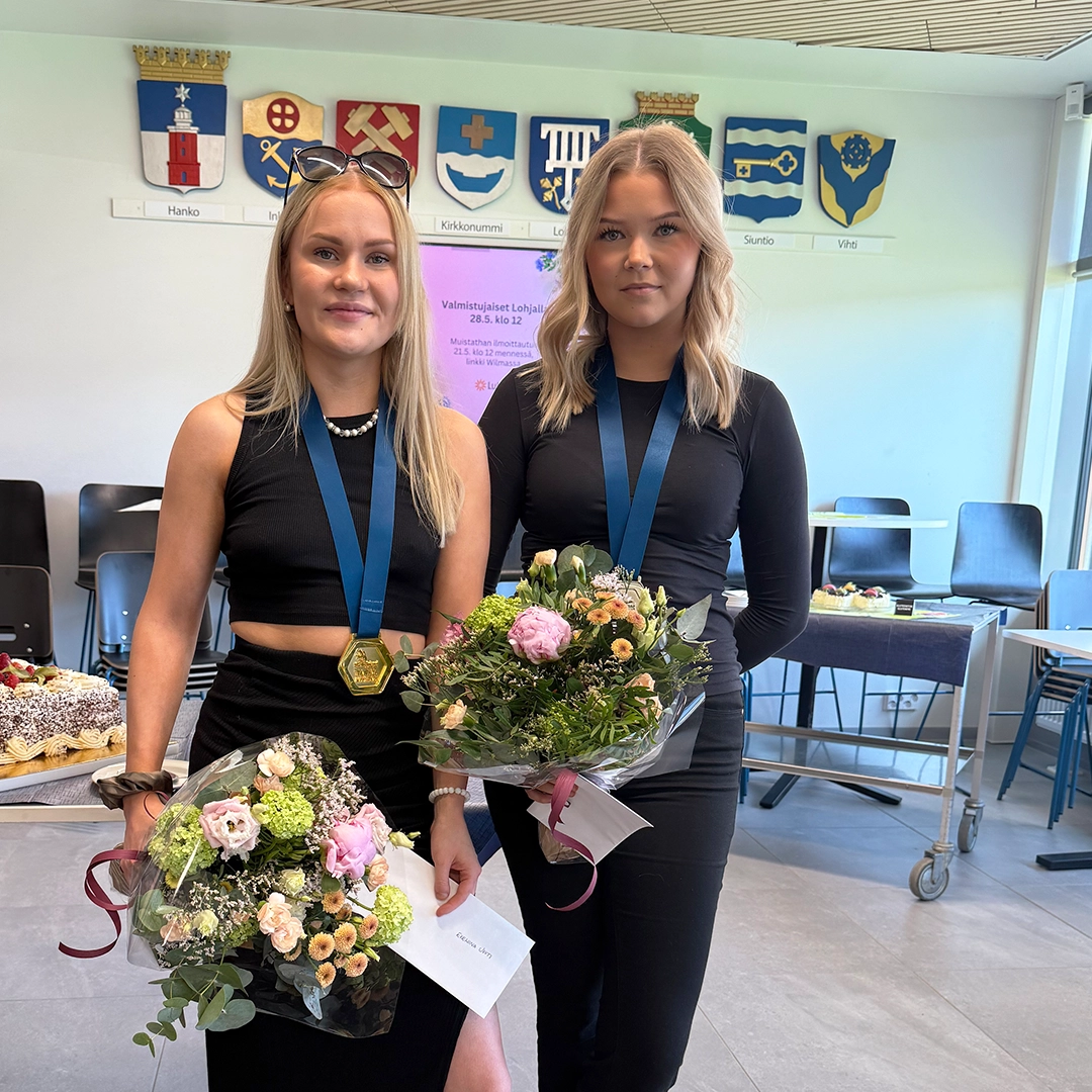 Kaksi vaaleahiuksista henkilöä mustissa vaatteissa, mitalit kaulassa ja värikäs kukkakimppu kädessä.