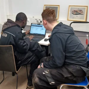 Kaksi henkilöä tummissa vaatteissa istuu ja katsoo työpöydällä olevaa päätettä.