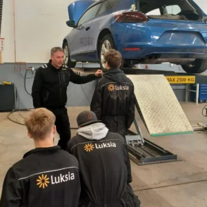 Neljä ihmistä autohuoltohallissa, jossa sininen auto.
