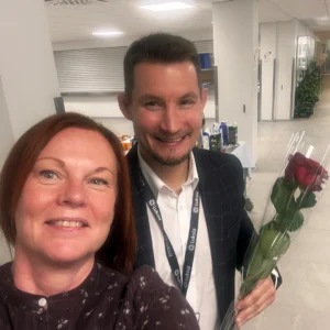 Kaksi henkilöä tummissa vaatteissa. Toisella henkilöllä on punainen ruusu kädessä.