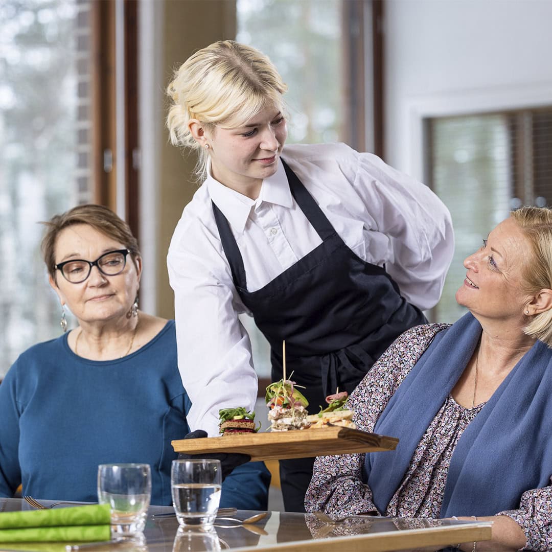 Kolme henkilöä ravintolassa. Kaksi asiakasta ja tarjoilija.