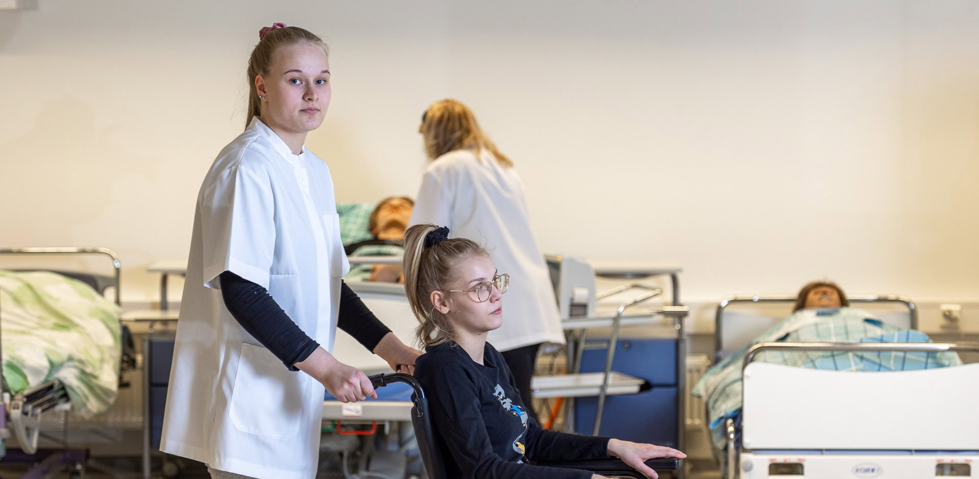 Henkilö valkoisessa asussa, työntää pyörätuolissa olevaa henkilöä, jolla on tummat vaatteet.