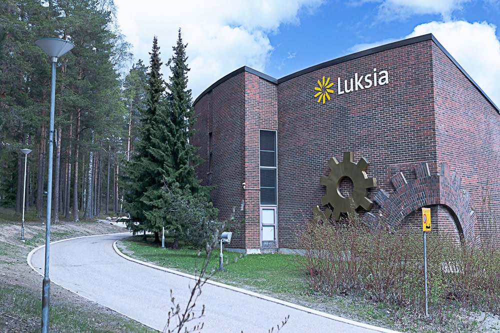 Ruskeatiilinen rakennus, sininen taivas ja vihreää metsää.