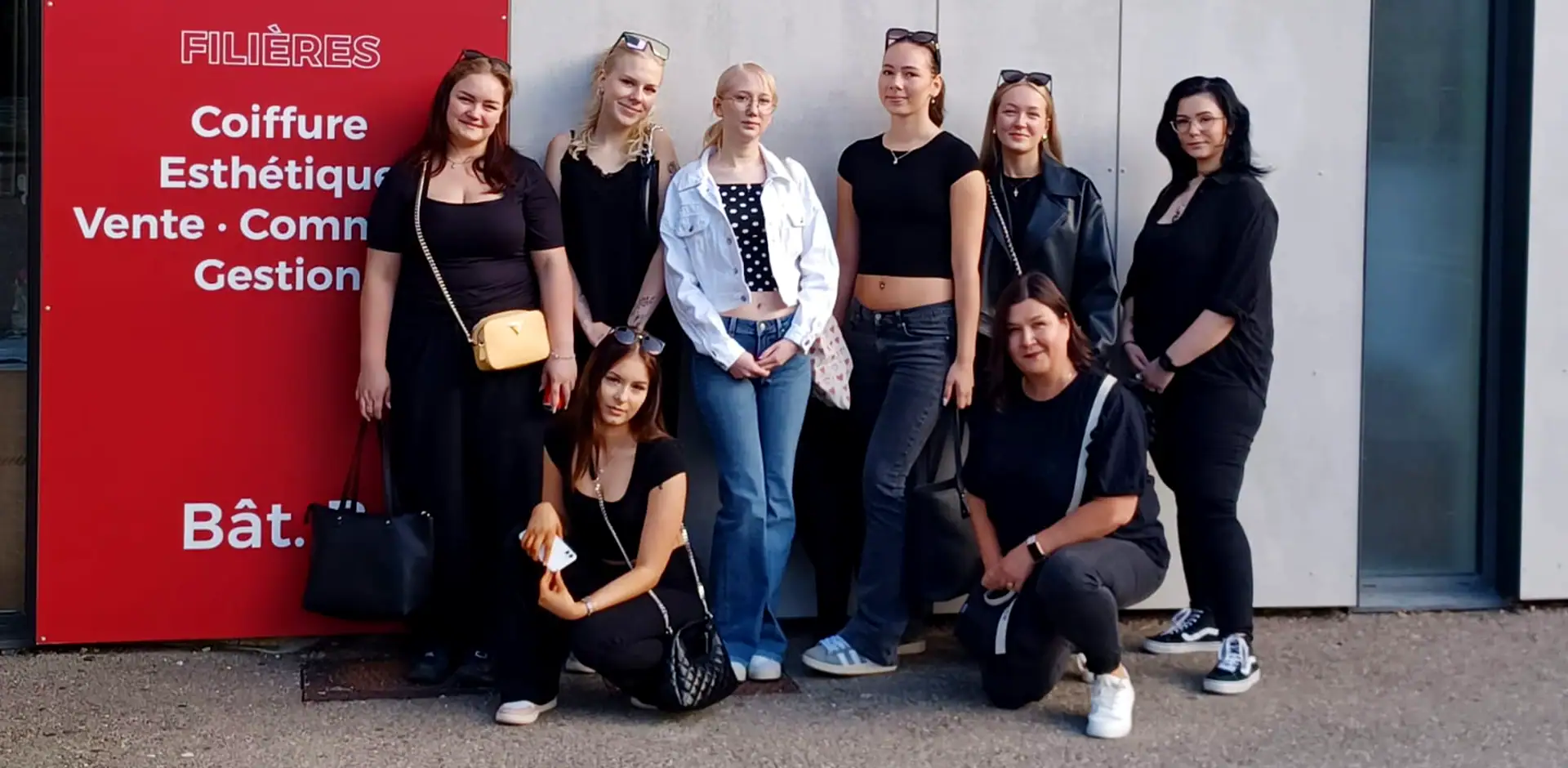 Ryhmä ihmisiä seisoo talon edustalla. Talon seinässä on punainen kyltti, jossa valokoista kirjoitusta.