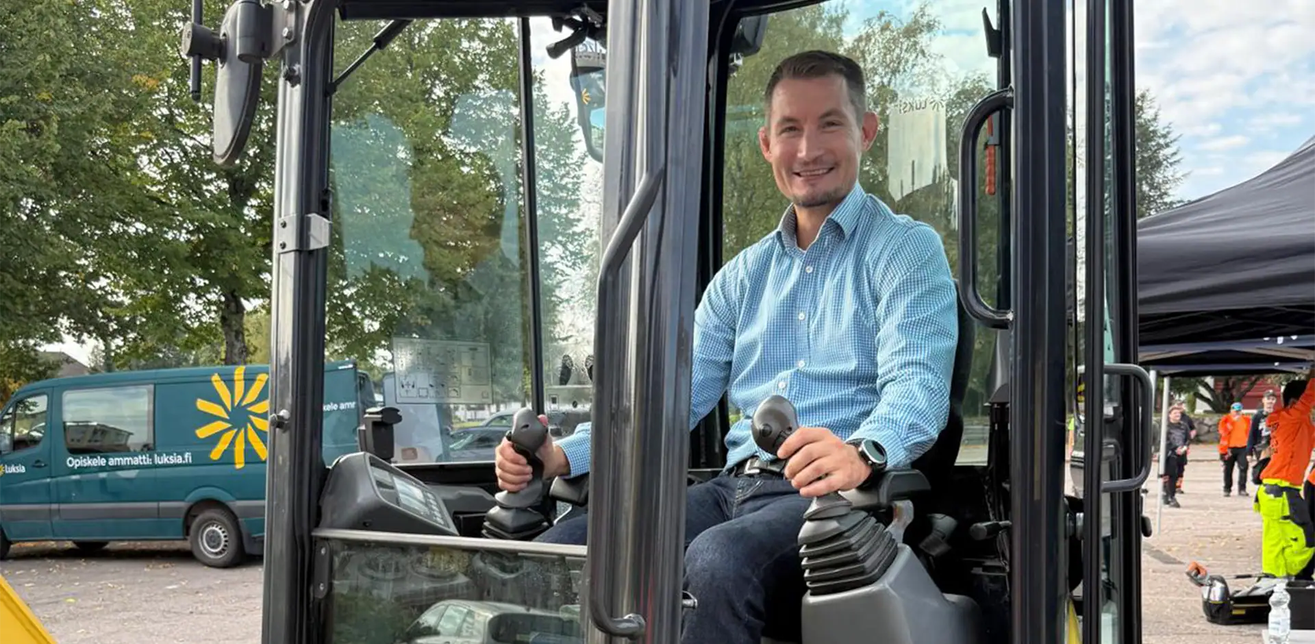 Henkilö sinisessä paidassa istuu keltaisen kaivinkoneen ohjaamossa.