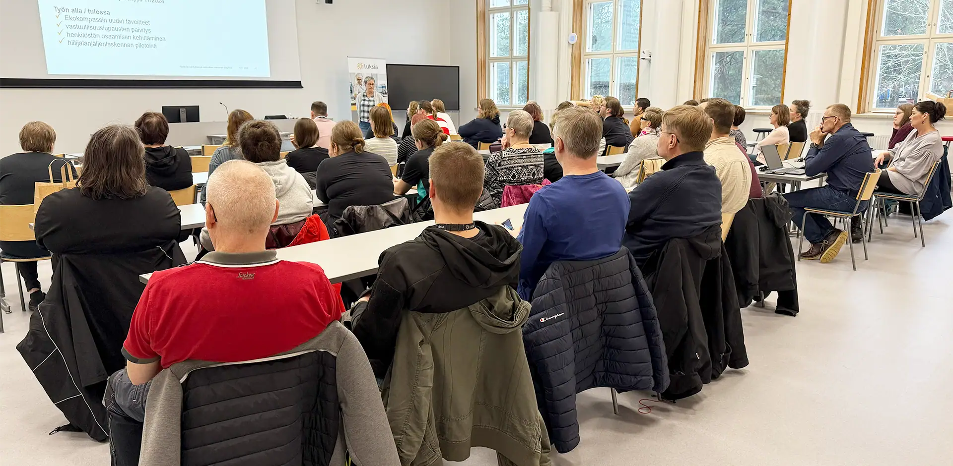 Luentosalissa joukko ihmisiä kuuntelee luennoitsijaa, joka seisoo yleisön edessä.