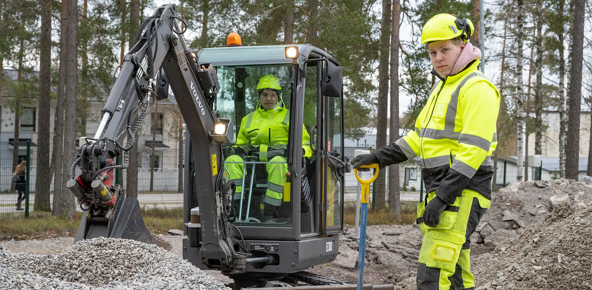 Kaivinkone ja kaksi rakentajaa työmaalla, työskentelemässä maaperän parissa.