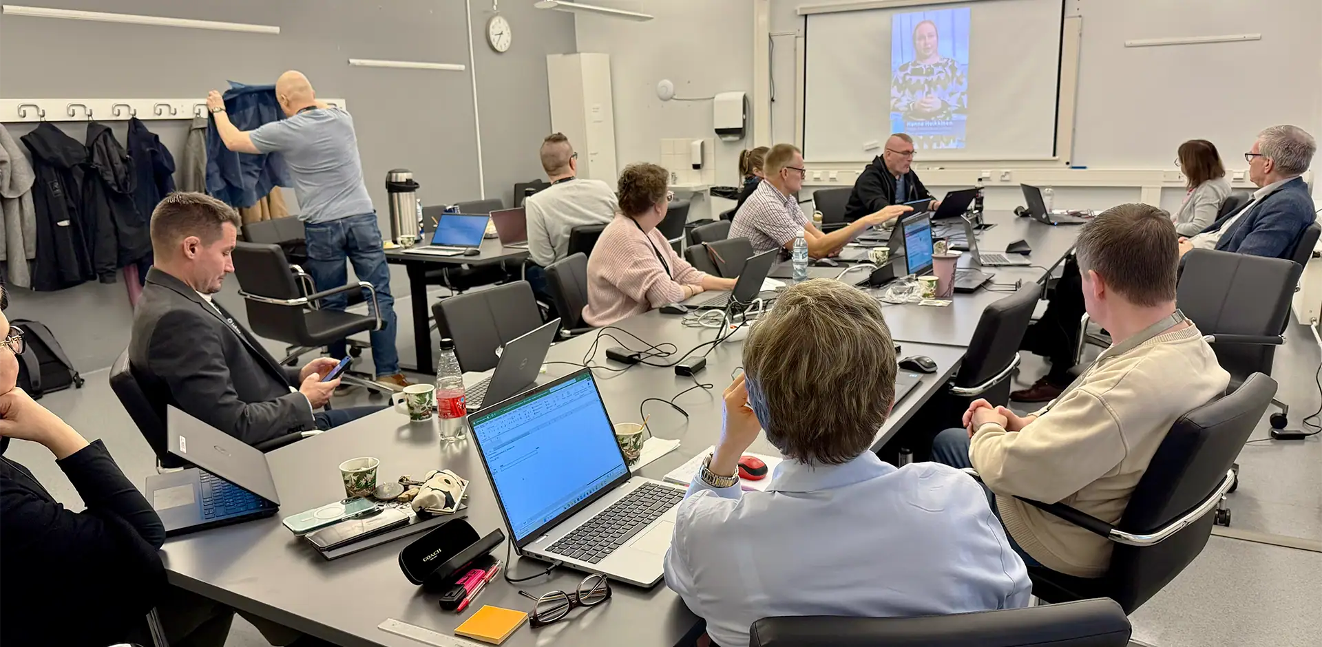 Ryhmä ihmisiä neuvotteluhuoneessa katsoo lähetystä, jossa henkilö seisoo ja puhuu.