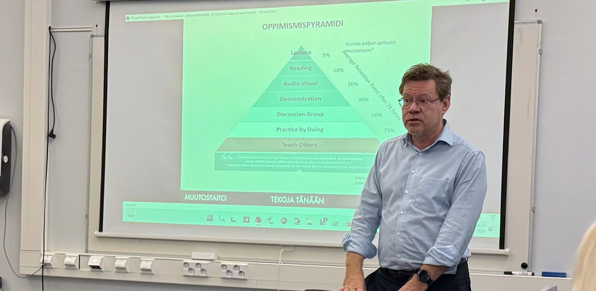 Henkilö vaaleassa paidassa seisoo luokan edessä ja pitää esitystä.
