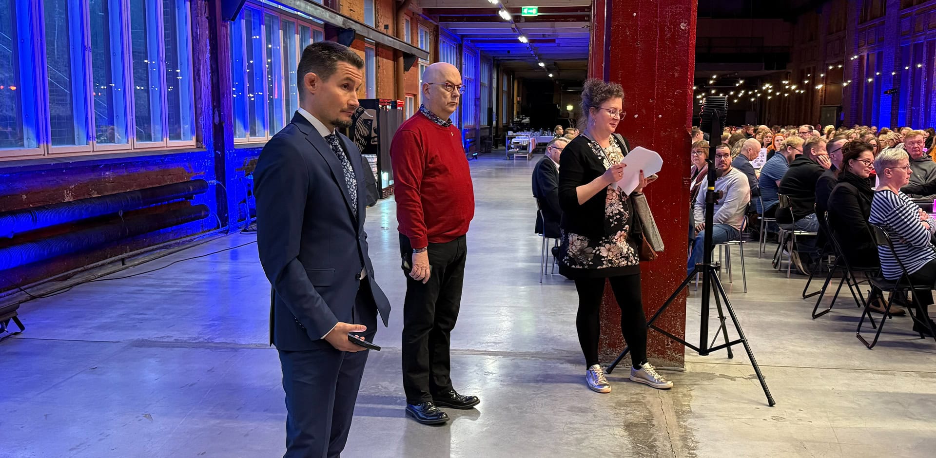 Kolme henkilöä seisoo ja katsoo kohti näyttämöä.