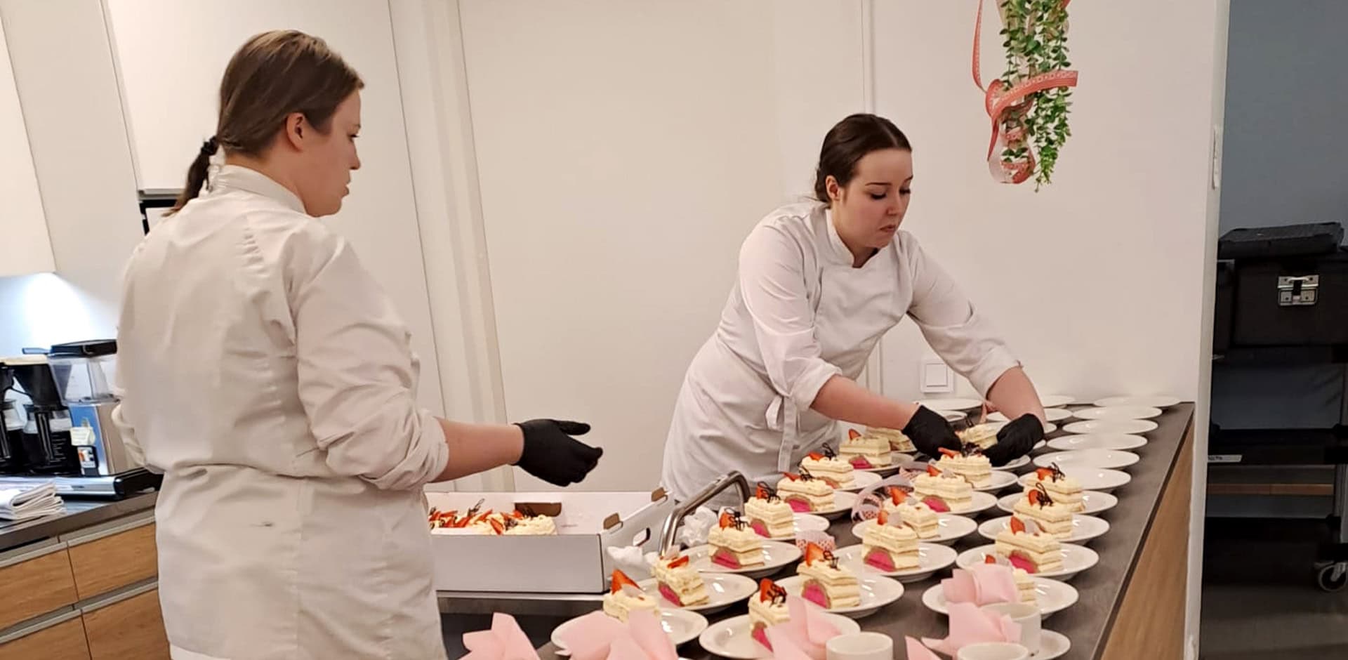 Kaksi henkilöä valkoisissa vaatteissa asettaa kakun paloja esille pöytään.